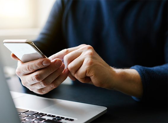 A person uses a smartphone while sitting at a desk with an open laptop.