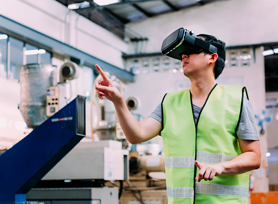A man in a neon vest uses VR goggles in an industrial setting, pointing forward. The environment is high-tech, suggesting innovation and focus.