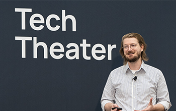 Speaker stands in front of Tech Theater sign on stage